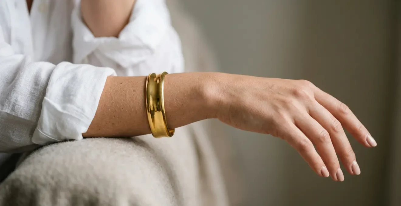 Close-up of a woman's wrist wearing a statement gold cuff bracelet against a neutral minimal outfit in natural light
