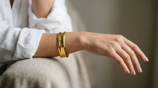 Close-up of a woman's wrist wearing a statement gold cuff bracelet against a neutral minimal outfit in natural light